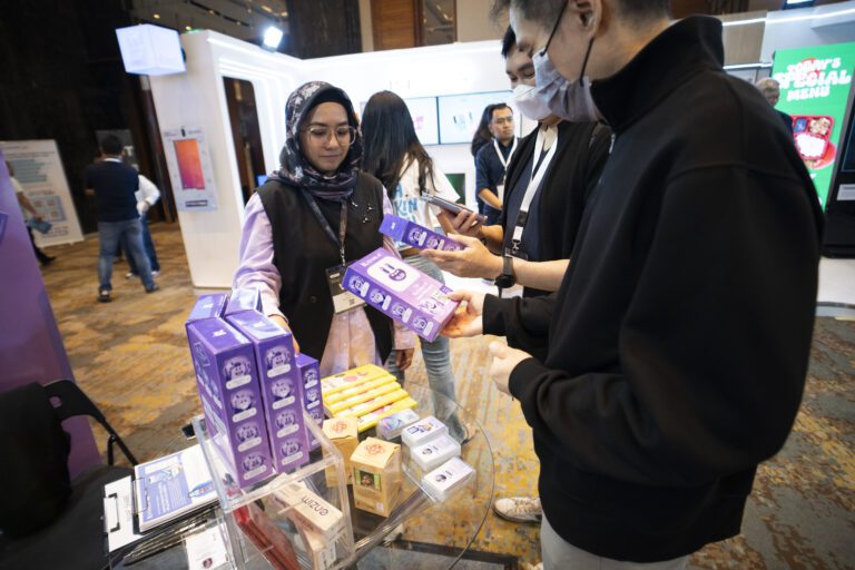 Woman wearing hijab and glasses examines products at a booth as a man in a mask offers packages from a display of purple boxes.