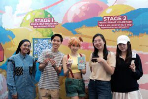 Group of five young adults posing for a photo in front of a colorful mural, some holding flyers and ice cream cones; everyone smiles at the camera.