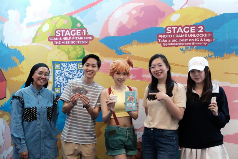 Group of five young adults posing for a photo in front of a colorful mural, some holding flyers and ice cream cones; everyone smiles at the camera.