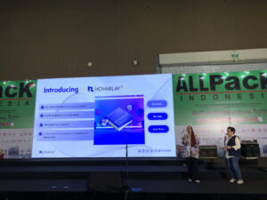 Stage at a trade show with a large screen reading 'Introducing R HOVARLAY', two presenters on the right, and a green ALLPACK banner in the background.