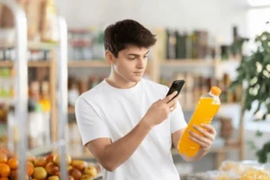 Man in a white t-shirt in a supermarket checks his phone while holding an orange juice bottle in his hand.