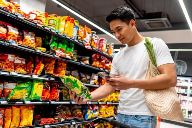 art99 - Hovarlay Man in a grocery store checks his phone while holding a bag of greens, with colorful snack shelves behind him.