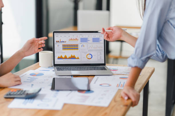 Two data analysts Working on data analysis dashboard for business strategy - Hovarlay Two people discuss data shown on a laptop displaying charts and graphs at a wooden table with reports and a calculator nearby in an office setting.