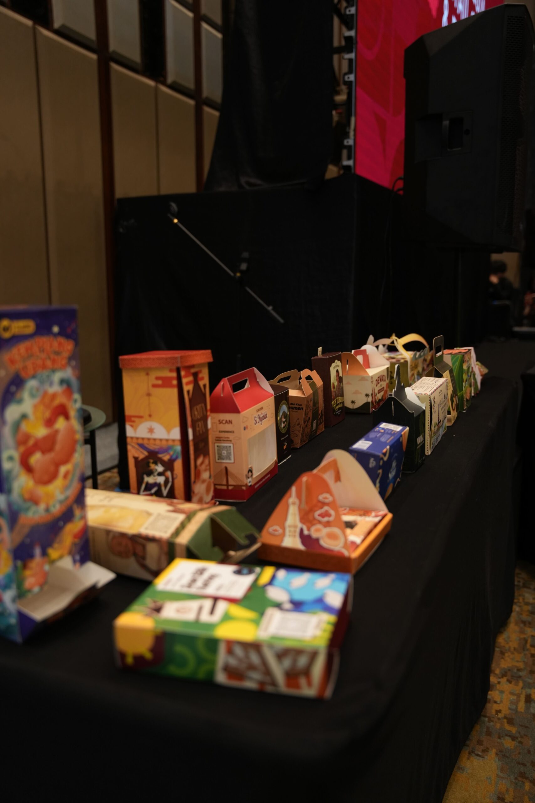 Row of colorful gift boxes arranged on a black table at an event, with a stage and red screen in the background.