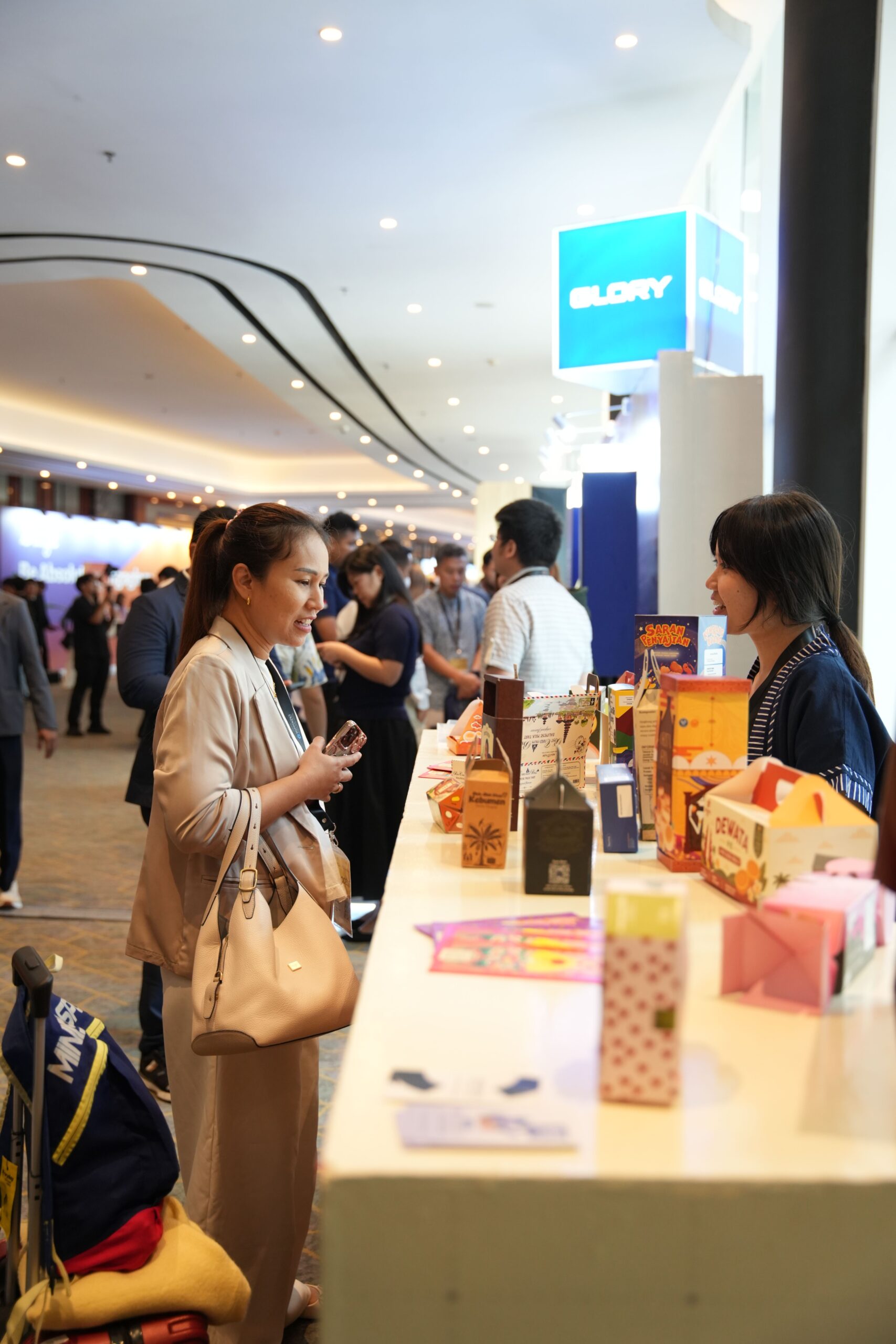 A woman in a beige coat browses boxed products laid out on a long white table at a busy indoor expo or trade show.