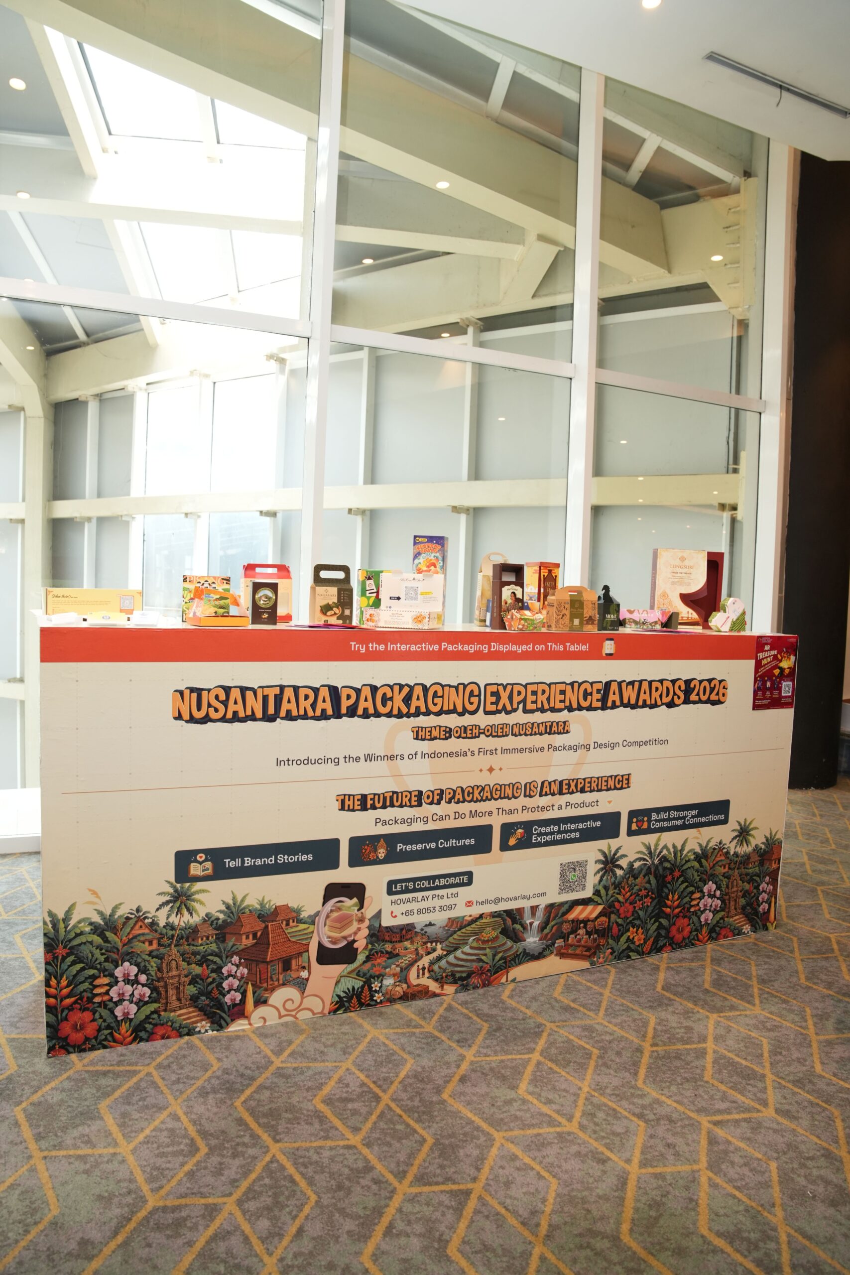 Exhibition booth displaying 'Nusantara Packaging Experience Awards 2026' banner with sample packaging boxes on a red counter above a tropical-themed backdrop.
