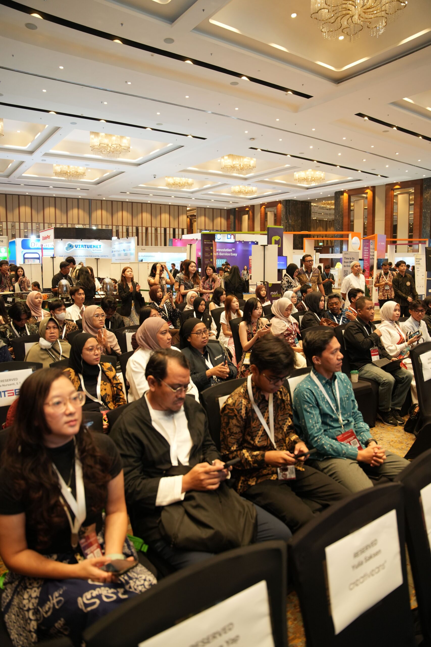 Crowded conference hall with rows of attendees and colorful exhibitor booths in the background, watching a presentation.