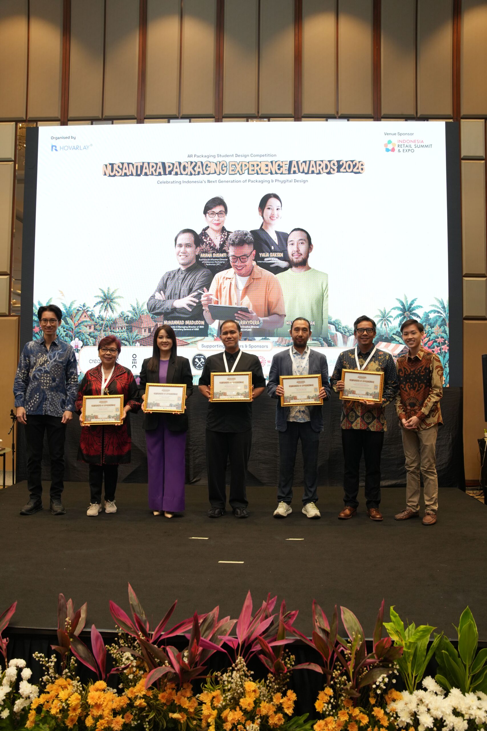 Eight people stand on a stage holding framed certificates, with a large event banner and vibrant flowers in front of them.