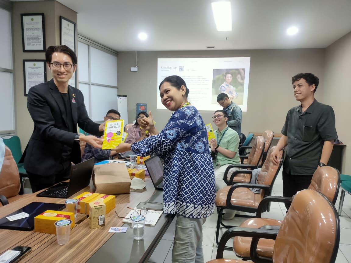 Man in a black suit hands a yellow boxed item to a smiling woman in a blue patterned blouse during a meeting in a conference room with colleagues present.