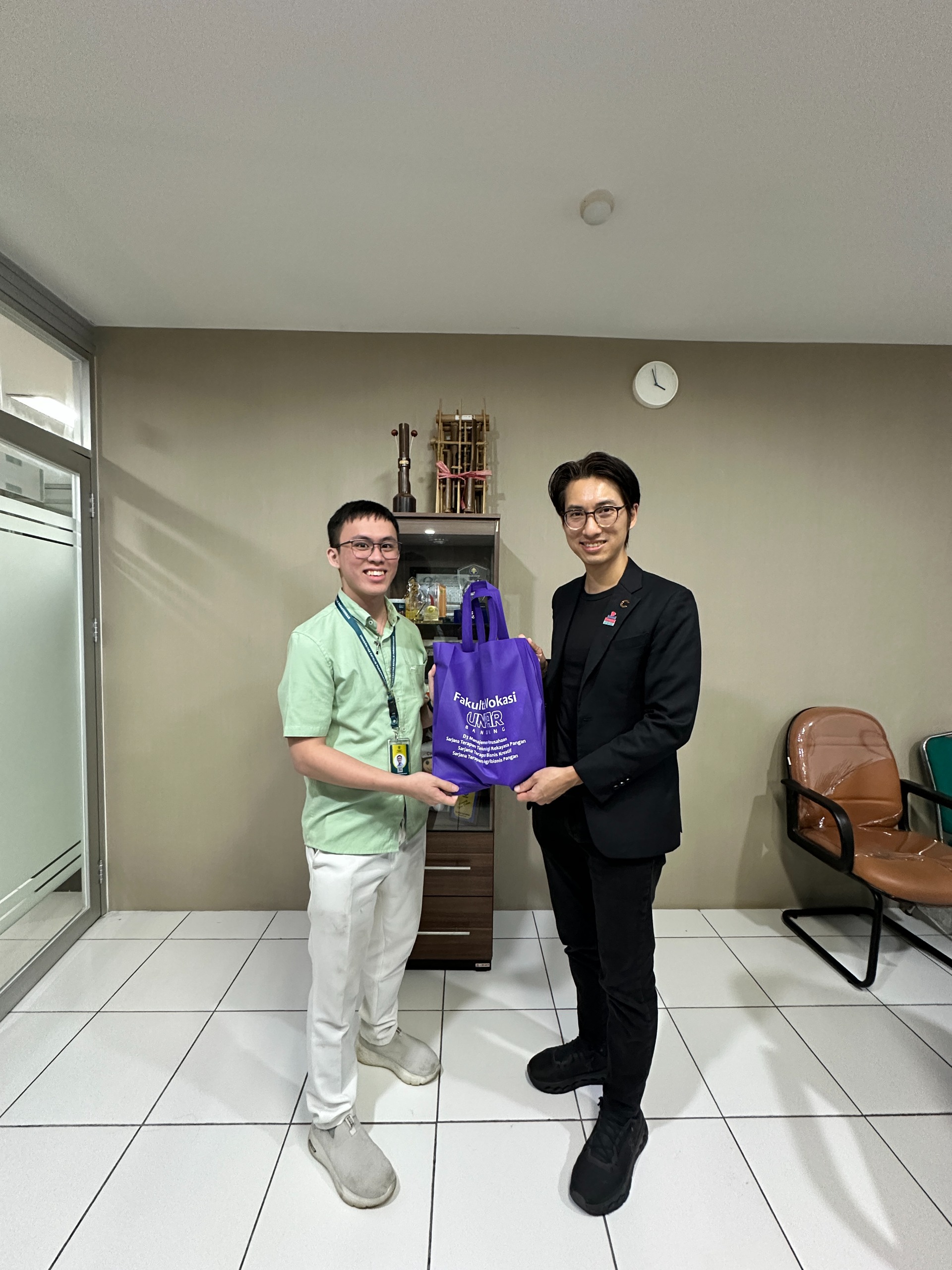 Two men in an office exchange a purple bag, standing side by side with a cabinet and clock behind them.