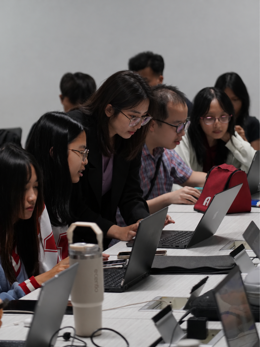 Group of students collaborating at a long table with laptops in a classroom environment.