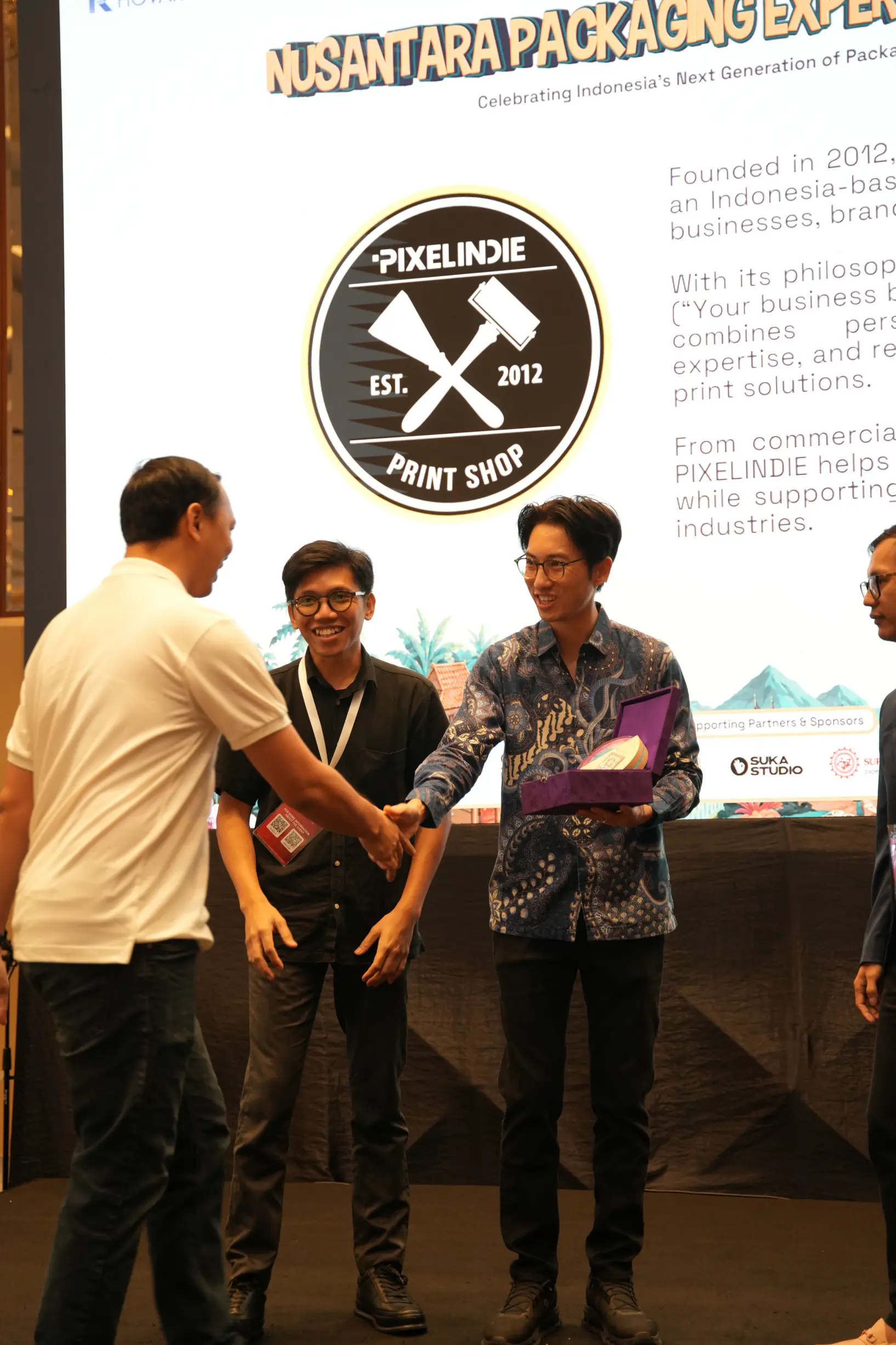 Group of men shaking hands on a stage with a large circular PixelIndie Print Shop logo in the background.