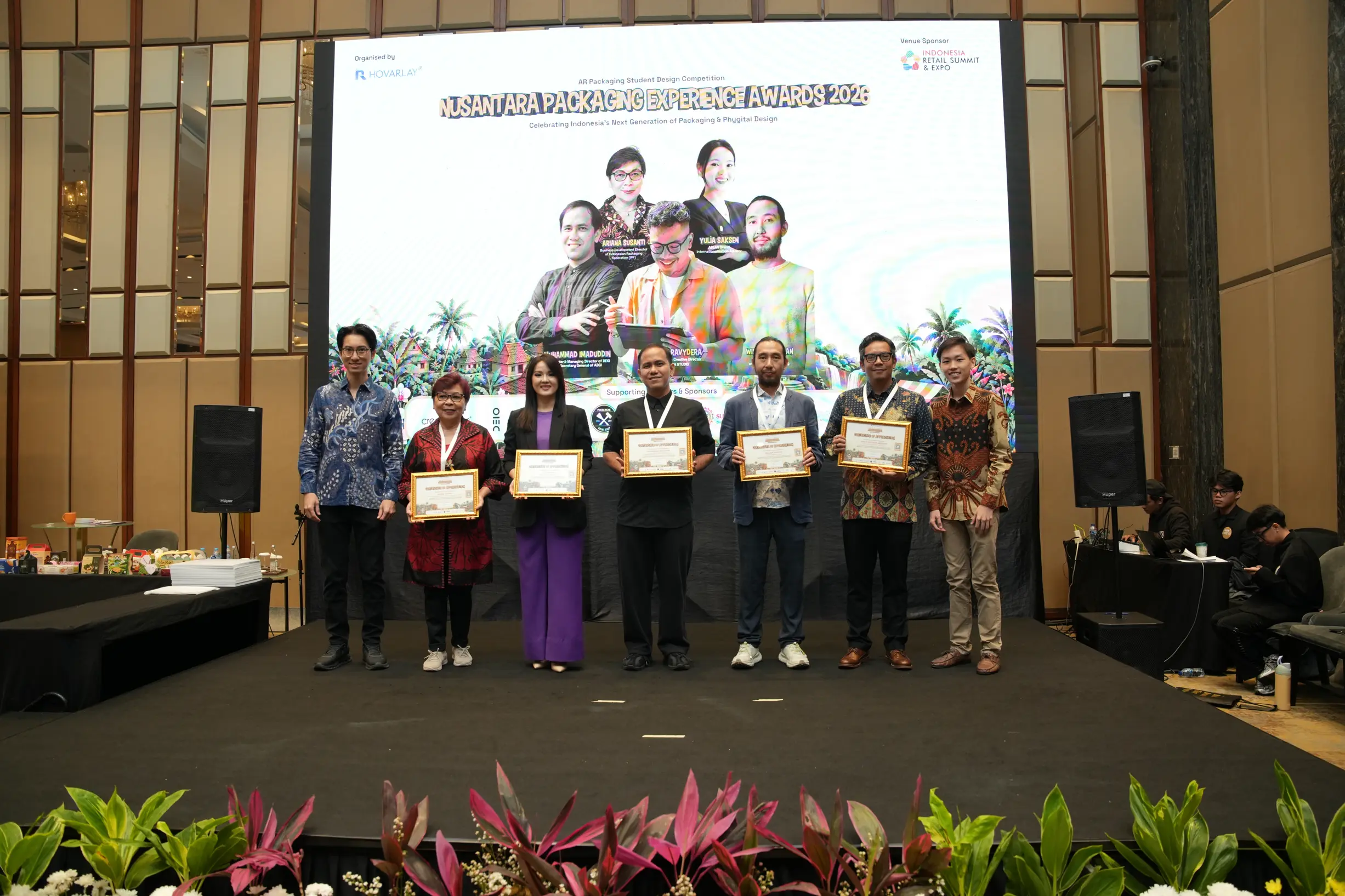 Group of eight people on a stage holding framed certificates at an awards ceremony, with a large poster backdrop and plants in the foreground.