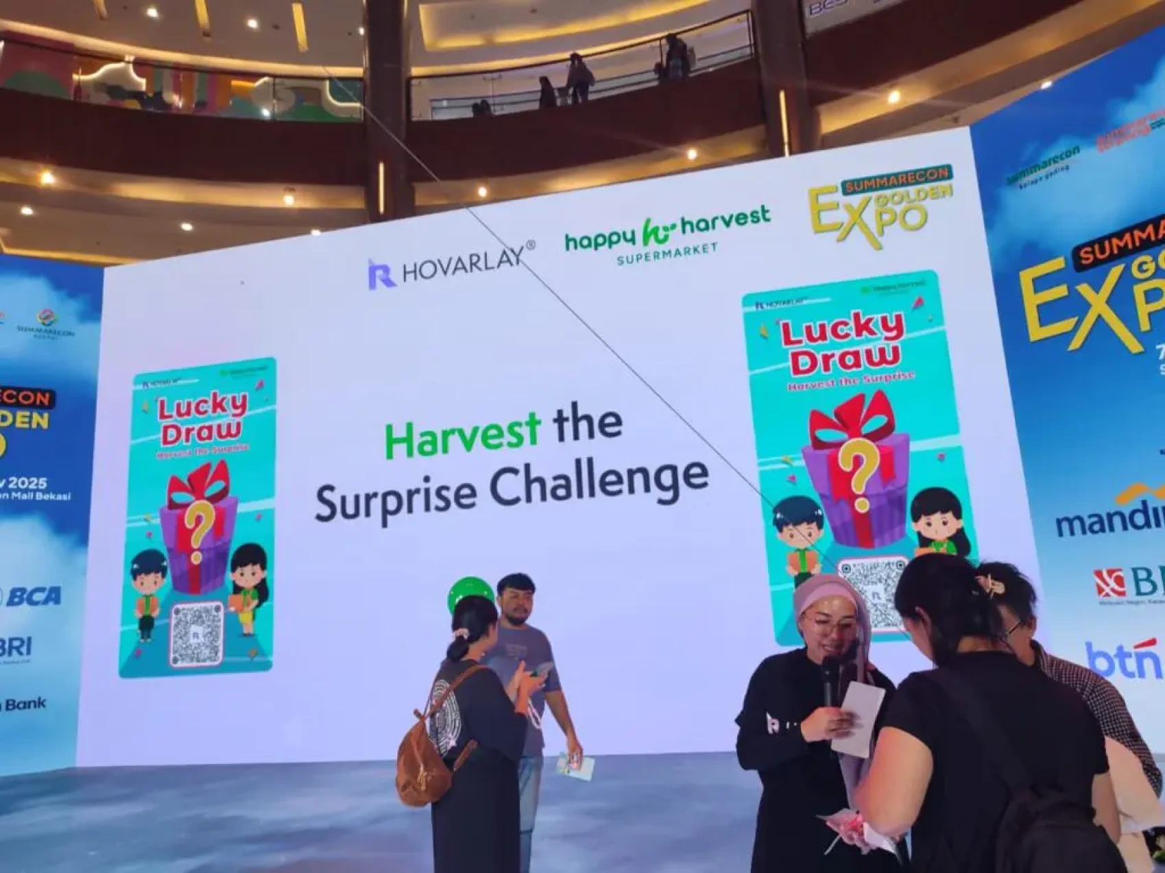 Stage backdrop reads 'Harvest the Surprise Challenge' with Lucky Draw posters and sponsor logos at a mall expo.
