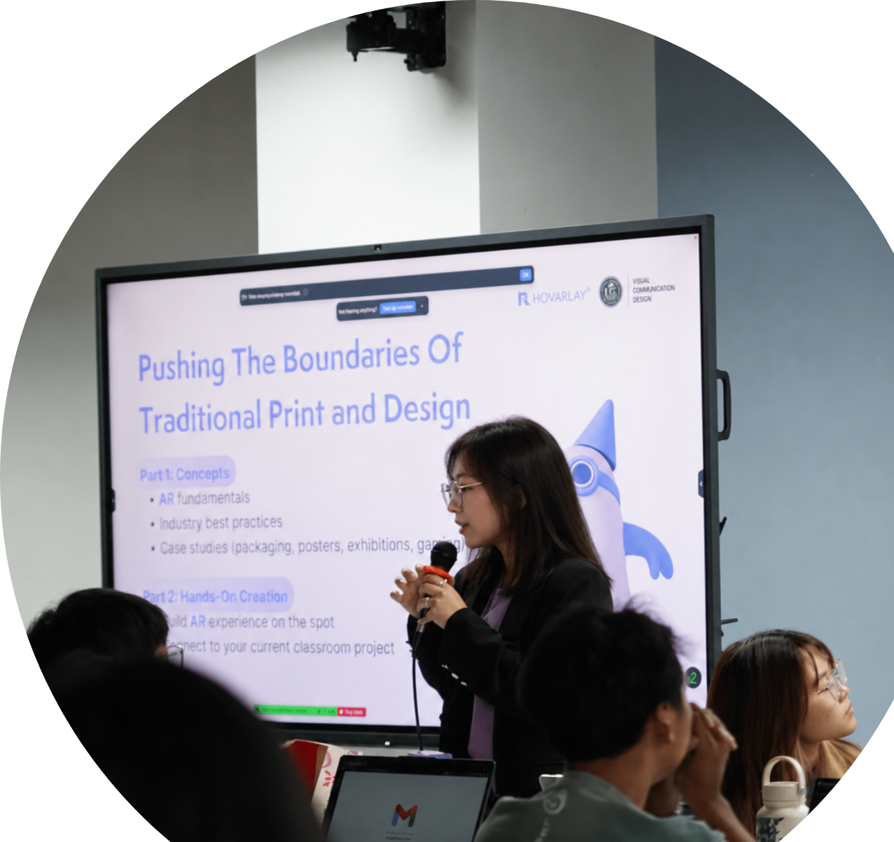 Female presenter speaks into a microphone beside a large screen displaying a slide titled 'Pushing The Boundaries Of Traditional Print and Design' in a conference room. Audience members are watching.